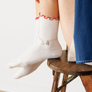 Person wearing a blue jacket and white socks with red trim and a silver charmed ankle bracelet, sitting on a wooden stool.