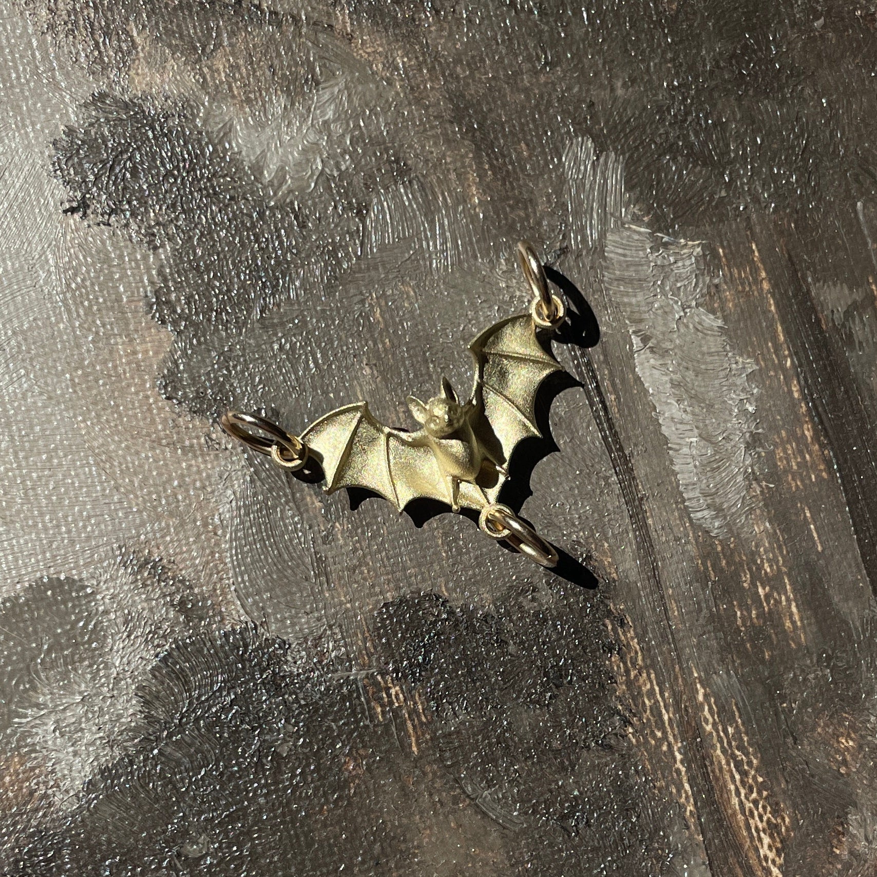 A close-up of a textured painting depicting a dark, moody forest scene with a small bat perched on a tree trunk. The artwork features a mix of dark gray, brown, and beige tones, with a small metallic bat-shaped ornament resting on the canvas.