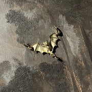 A close-up of a textured painting depicting a dark, moody forest scene with a small bat perched on a tree trunk. The artwork features a mix of dark gray, brown, and beige tones, with a small metallic bat-shaped ornament resting on the canvas.