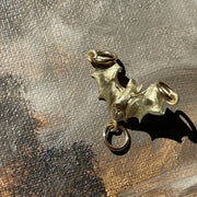 A small gold-toned metal charm shaped like a stylized leaf or winged motif with two loops, photographed on a textured brown and gray surface with strong sunlight and shadow.
