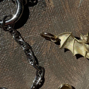 A close-up of a dark metallic chain necklace with a bat-shaped pendant, displayed on a textured brown surface next to a small gold-toned bead or charm.