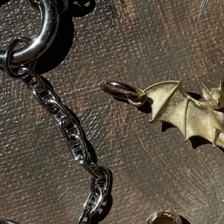 A close-up of a dark metallic chain necklace with a bat-shaped pendant, displayed on a textured brown surface next to a small gold-toned bead or charm.