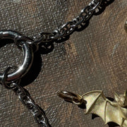 A close-up of a dark metallic chain necklace with a bat-shaped pendant, displayed on a textured brown surface next to a small gold-toned bead or charm.
