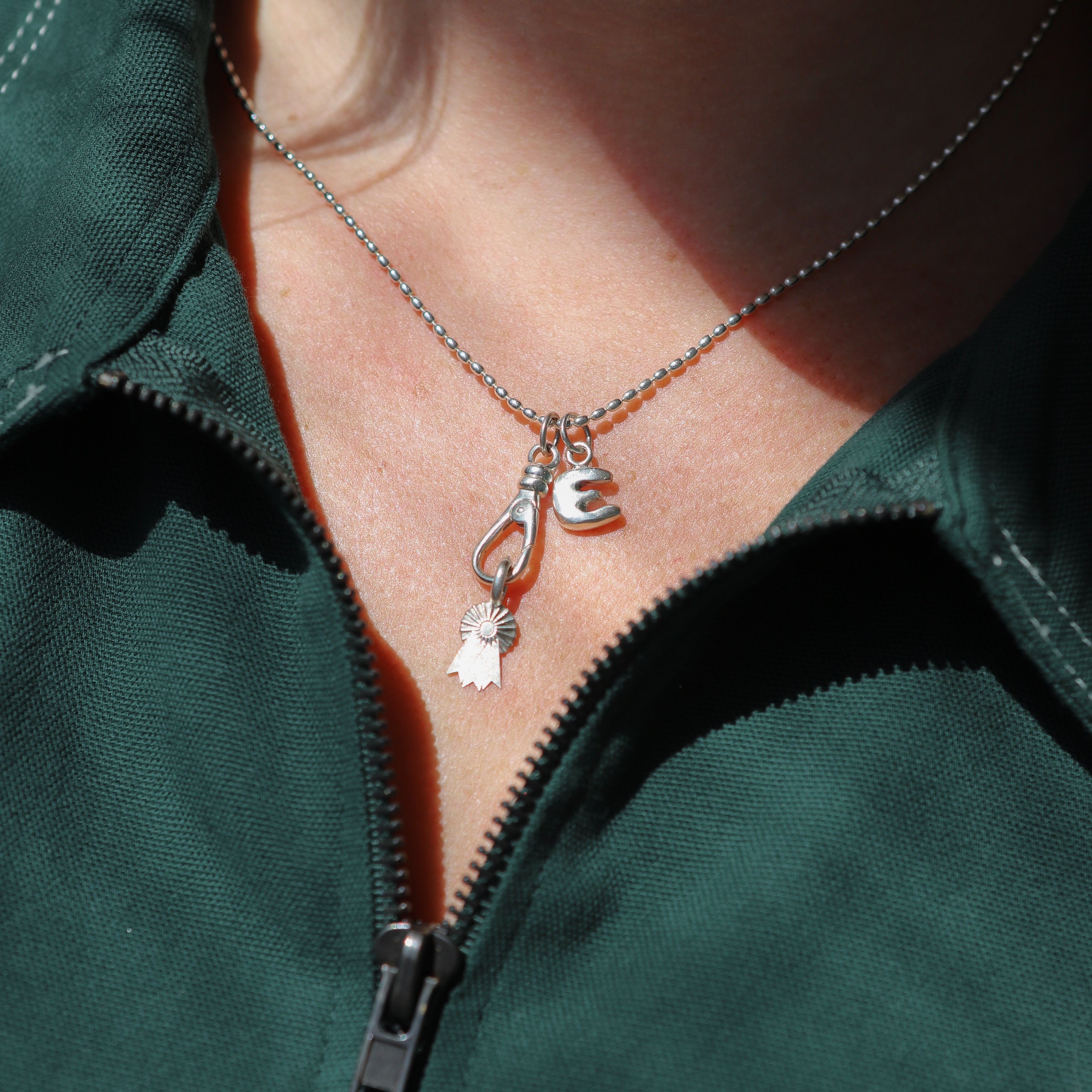 Close-up of a person wearing a green jacket with a necklace featuring a 'E' and heart pendant.