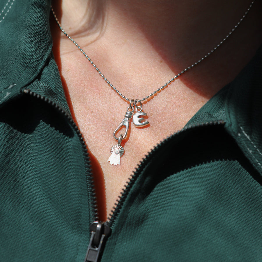 Close-up of a person wearing a green jacket with a necklace featuring a 'E' and heart pendant.