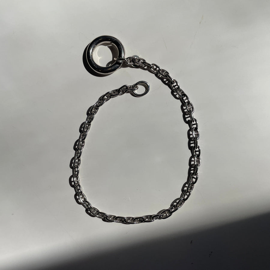 A sterling silver metal chain bracelet with a textured, rope-like link pattern and a circular ring charm, displayed on a reflective gray surface.