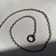 A sterling silver chain necklace with a matching circular ring clasp, coiled on a light gray surface with harsh direct sunlight casting shadows.