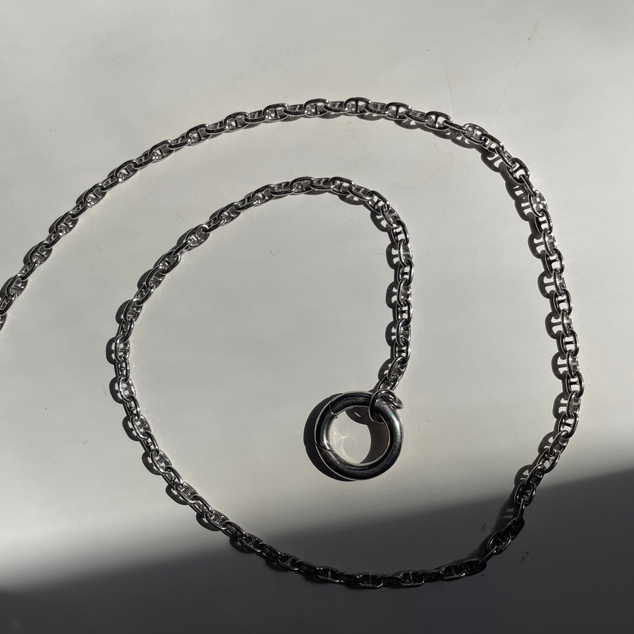 A sterling silver chain necklace with a matching circular ring clasp, coiled on a light gray surface with harsh direct sunlight casting shadows.