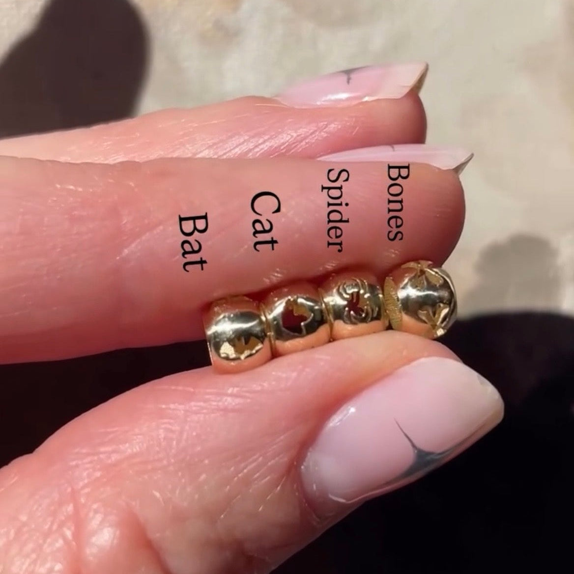 A close-up of a hand holding a small gold spacer beads with four cat-themed bead charms labeled 'Bat', 'Cat', 'Spider', and 'Bones'.